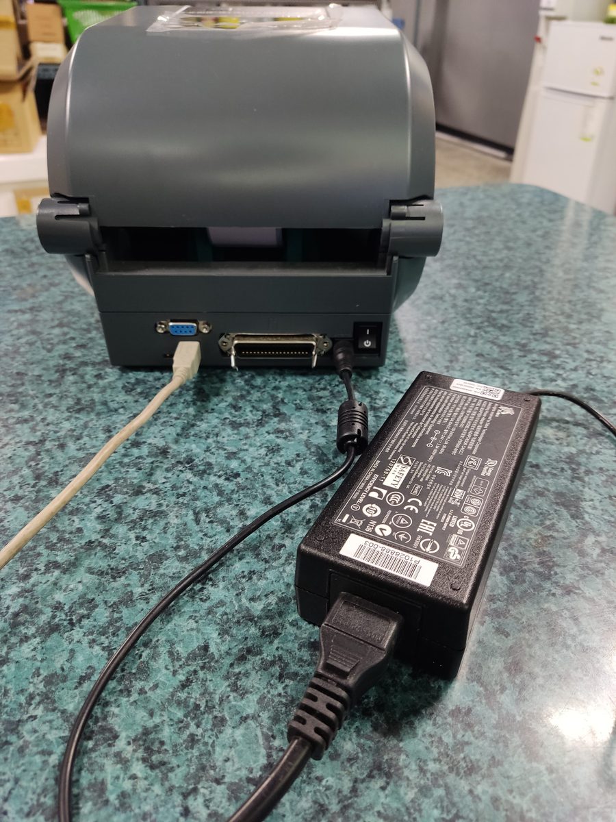 제브라 데스크톱 프린터 뒷면의 시리얼·패러럴·USB 단자 옆에 전원 어댑터 케이블을 꽂아 둔 모습